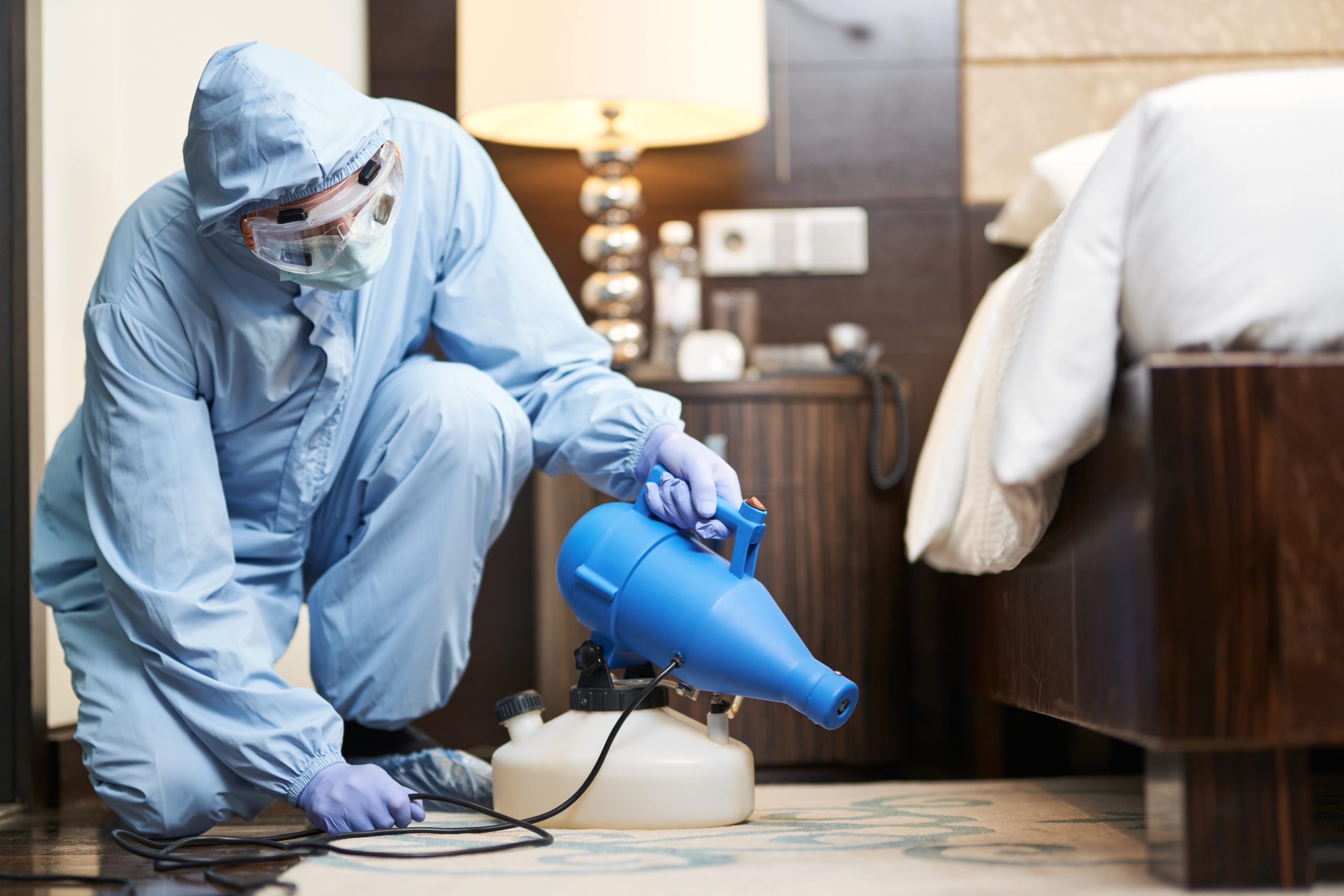 Technician in protective gear performing bed bugs pest control near a bed