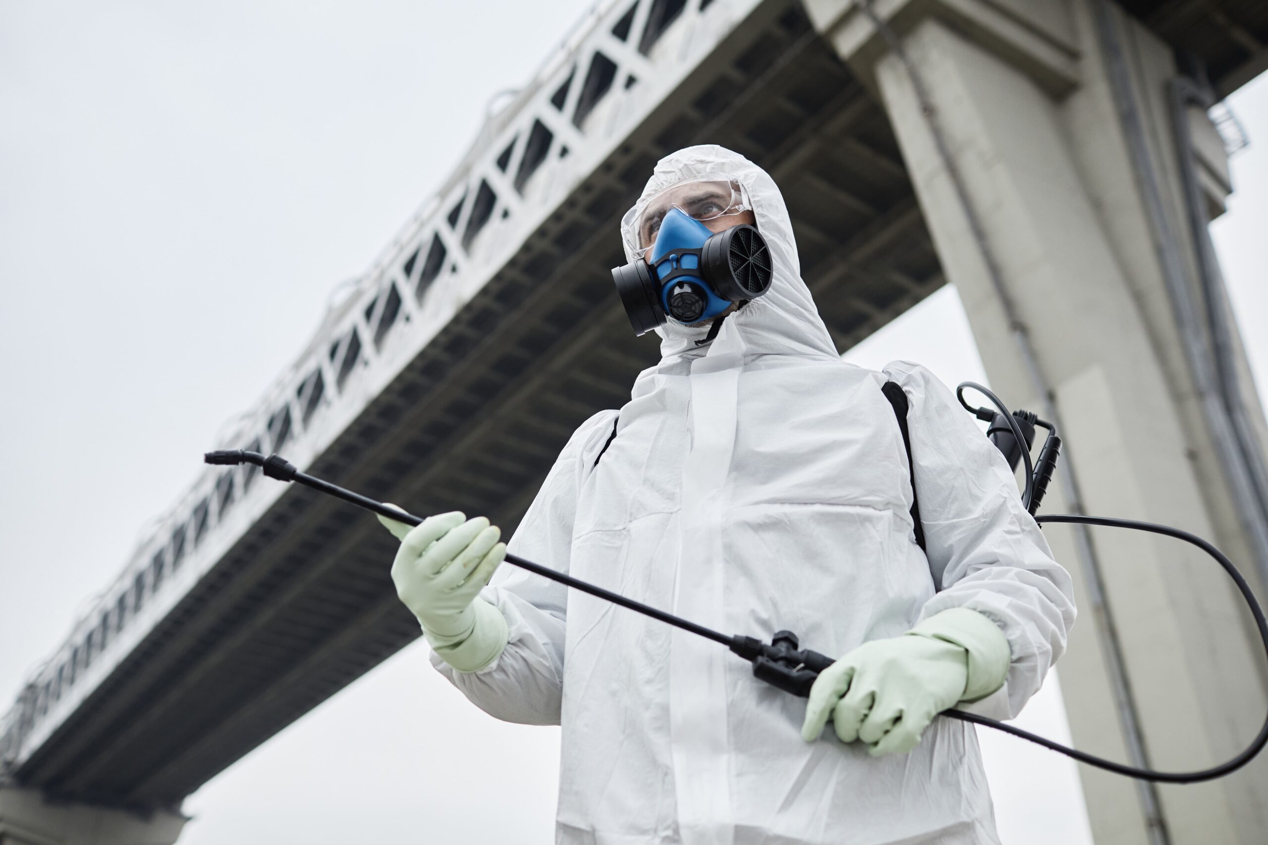 Commercial pest control service worker wearing full protective gear, standing outdoors near a bridge and holding spraying equipment.